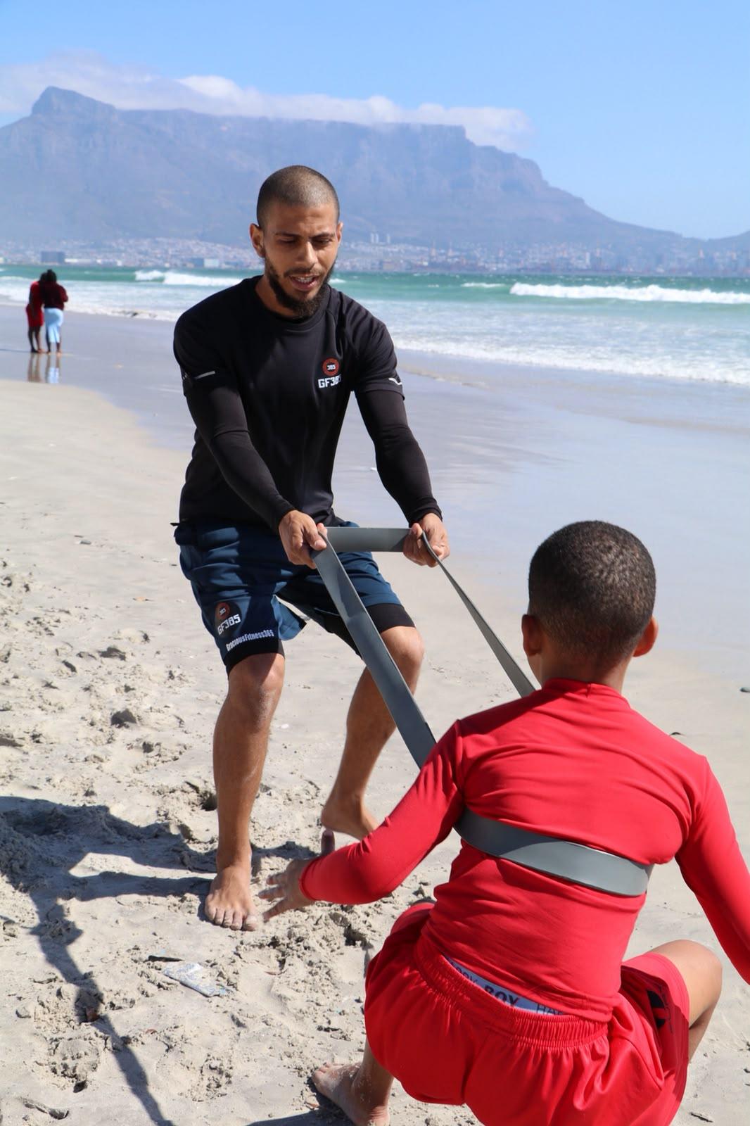 Gracious holding a resistance band while an athlete drives forward on the beach